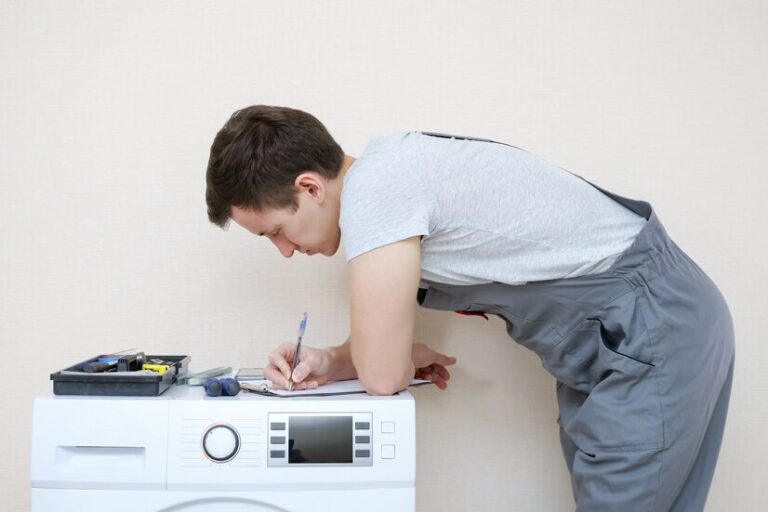young serviceman grey uniform with set tools writes clipboard examining modern washing machine with screen control panel room